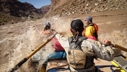 A guide rows an oar raft through a rapid on the Colorado River as passengers in the front are splashed by the waves.